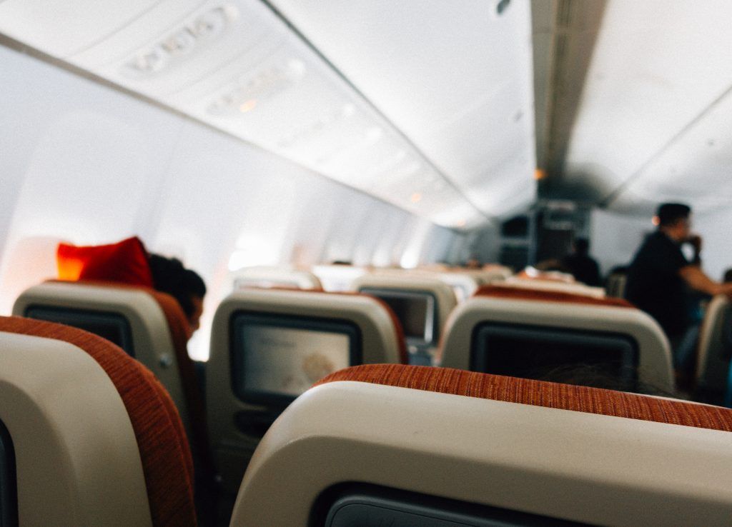 Ein Flugzeug-Kabineninneres mit Sitzreihen, jeder mit einem kleinen Bildschirm. Die Sitze sind beige und haben orangefarbene Besätze. Die Kabine ist gut beleuchtet.