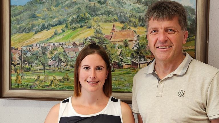 Ein lächelnder Mann und eine Frau stehen vor einem Landschaftsbild mit einem Dorf und Hügeln. Sie wirken beide fröhlich.