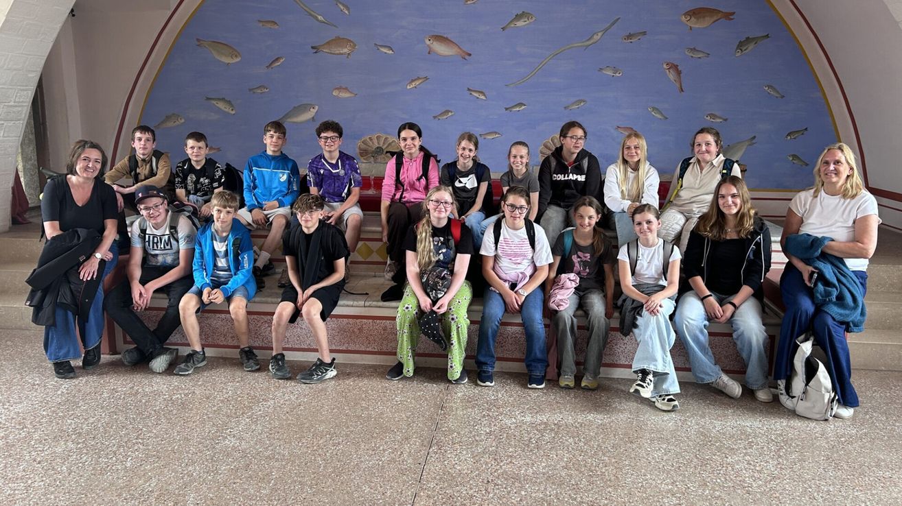 Eine Gruppe von Schülern sitzt auf einer Bank vor einer Wandmalerei mit Fischen. Sie tragen Freizeitkleidung und scheinen für ein Foto zu posieren.