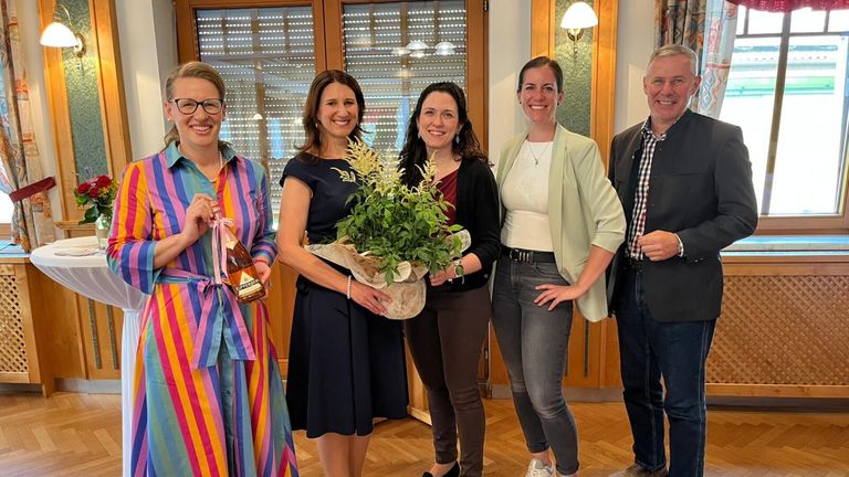 Fünf Menschen stehen in einem Raum zusammen. Die Frau in der Mitte hält einen Blumenstrauß. Eine andere Frau auf der linken Seite hält eine Flasche. Sie alle lächeln.