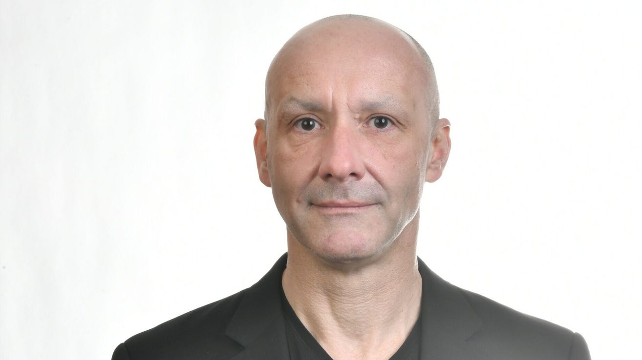 Ein Mann mit kahlem Kopf, der ein schwarzes Hemd trägt, lächelt und scheint für ein Foto zu posieren.