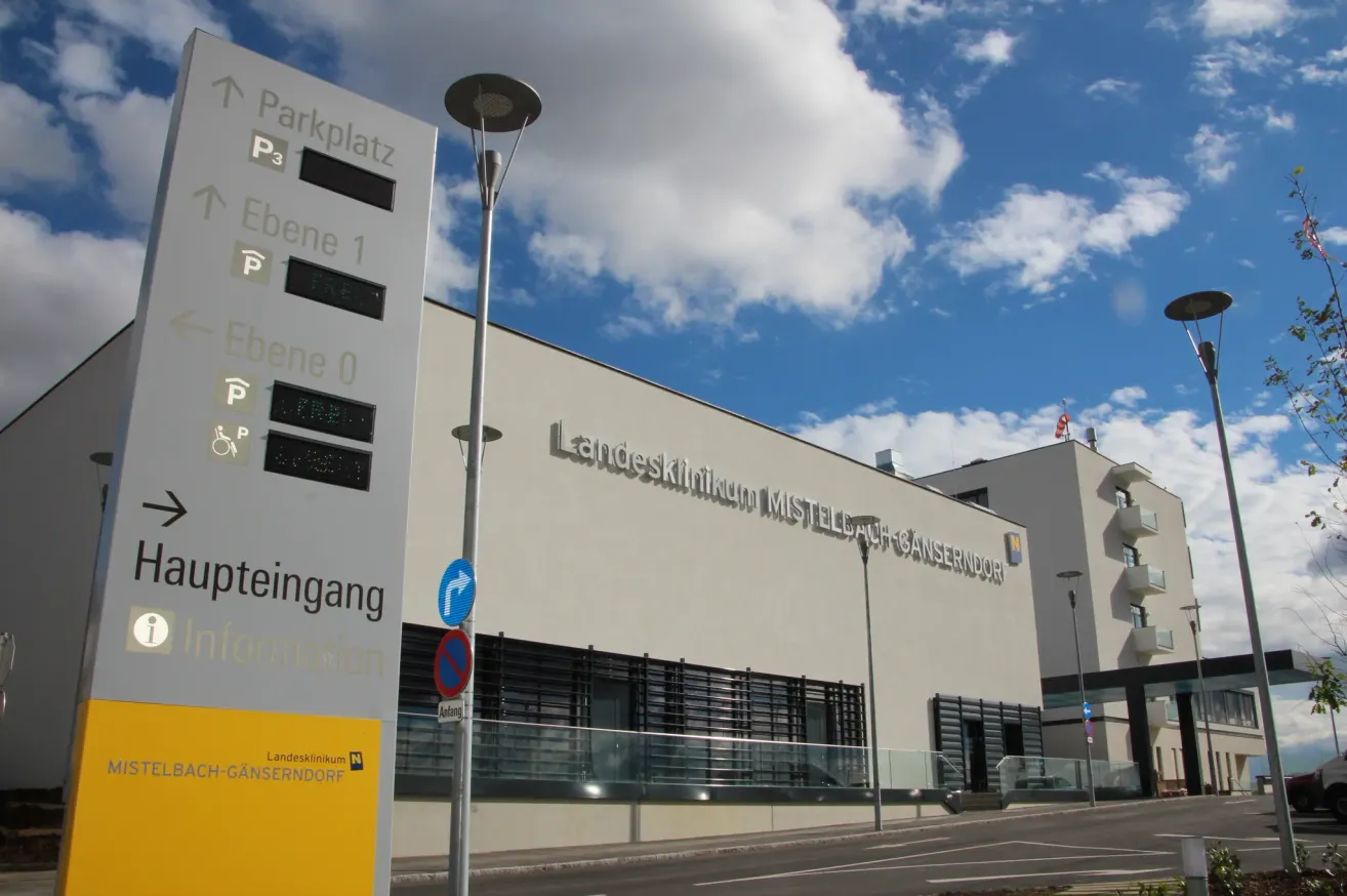 Ein großes weißes Gebäude namens Landesklinikum Mistelbach-Gänserndorf steht unter einem teilweise bewölkten Himmel. Es hat Glasfenster und ein Parkuhrschild in der Nähe.