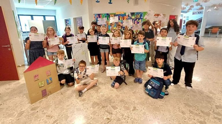 Ein Klassenzimmer voller Schüler, die Urkunden halten, einige sitzen, andere stehen, mit einem Schüler, der eine Zeichnung hält.