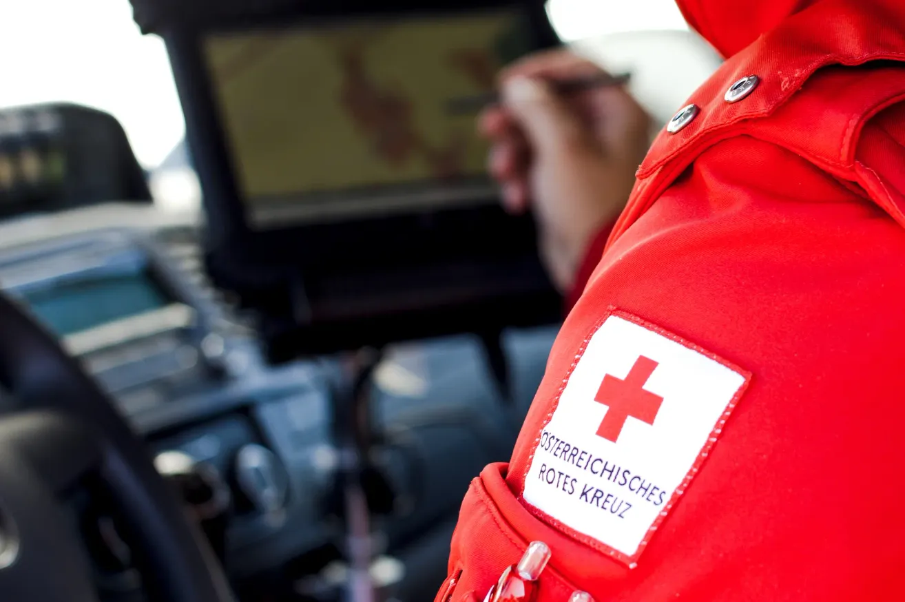 Eine Person in einem roten Jackett mit einem roten Kreuz-Patch arbeitet an einem Computer. Das Jackett hat eine verwackelte Hand und einen Computerbildschirm im Hintergrund.