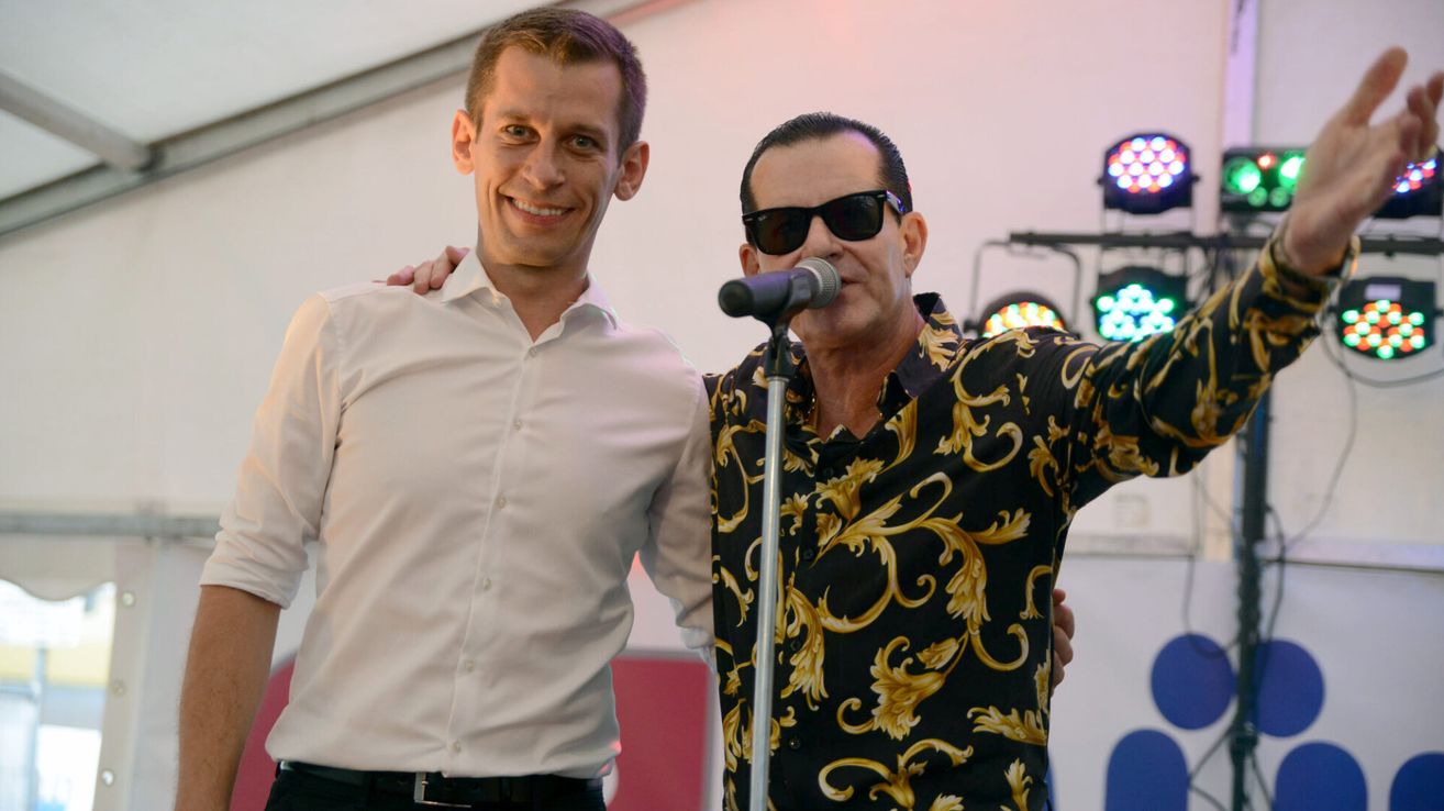 Zwei Männer posieren für ein Foto, einer in einem weißen Hemd und der andere in einem schwarz-golden gemusterten Hemd. Der Mann im gemusterten Hemd singt in ein Mikrofon.