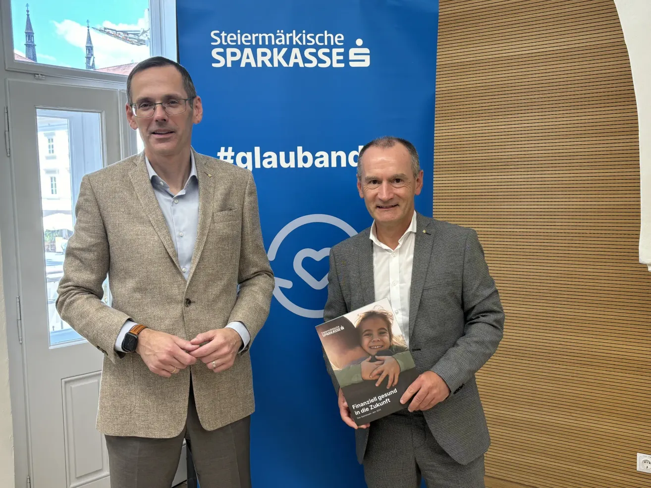 Zwei Männer stehen nebeneinander vor einem blauen Banner mit der Aufschrift 'Stmk Sparkasse'. Ein Mann hält ein Buch und der andere lächelt.