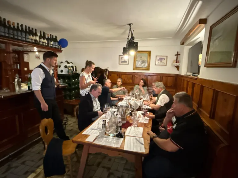 A group of people sit around a table in a restaurant, engaged in conversation. A man in a white shirt stands near a bar with wine bottles. A server stands at the head of the table.