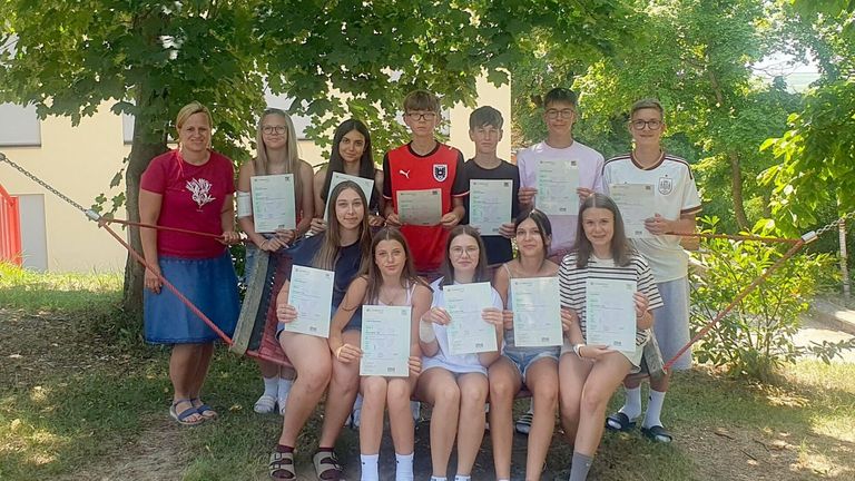 Eine Gruppe von Schülern posiert für ein Foto mit Urkunden vor einem Baum. Sie tragen alle Freizeitkleidung und lächeln. Einige tragen eine Brille, und ein Gebäude ist im Hintergrund.