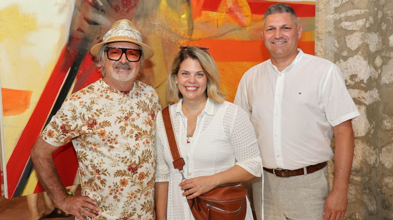 Three people stand before a colorful abstract painting, smiling. The man on the left wears a floral shirt and hat. The woman in the middle has a necklace and a brown bag. The man on the right wears a white shirt and pants.