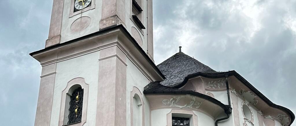 Ein hoher Kirchturm mit einer Uhr, einem Dachreiter und einem Fenster mit kunstvoller Eisenarbeit steht unter einem bewölkten Himmel.