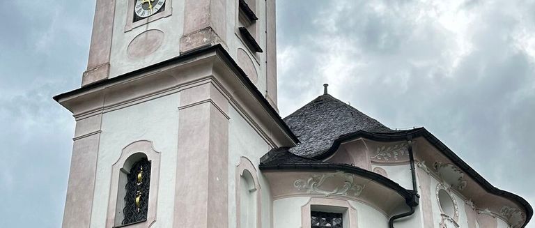 Ein hoher Kirchturm mit einer Uhr, einem Dachreiter und einem Fenster mit kunstvoller Eisenarbeit steht unter einem bewölkten Himmel.