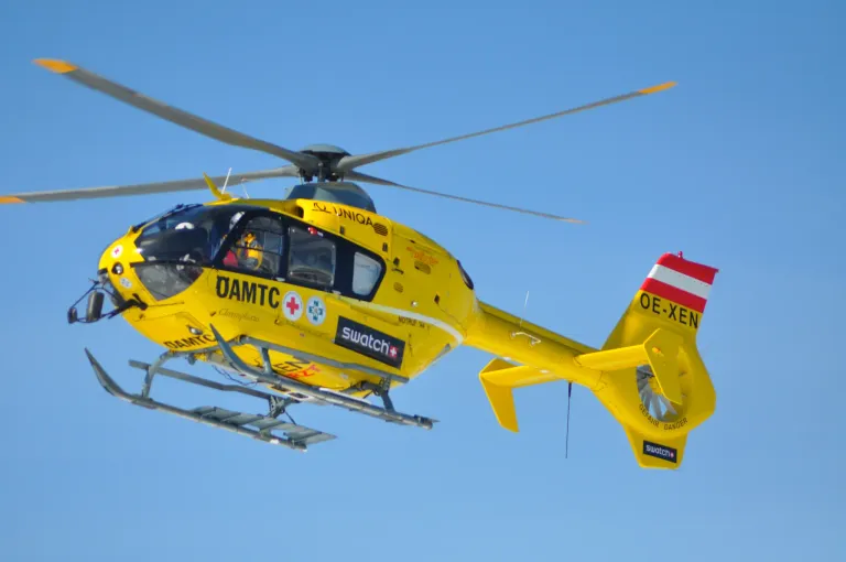 Ein gelber Hubschrauber mit 'OAMTC' und 'Swatch' auf der Seite fliegt am Himmel. Am Heck steht 'OE-XEN'. Er hat ein rotes Kreuz-Symbol und eine Notfallnummer.