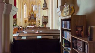 Eine Reihe von Kirchenbänken in einer Kirche, mit einem Altar und einem Bücherregal mit Büchern und einem Schild, das 'Generationentreff Bibliothek' steht.