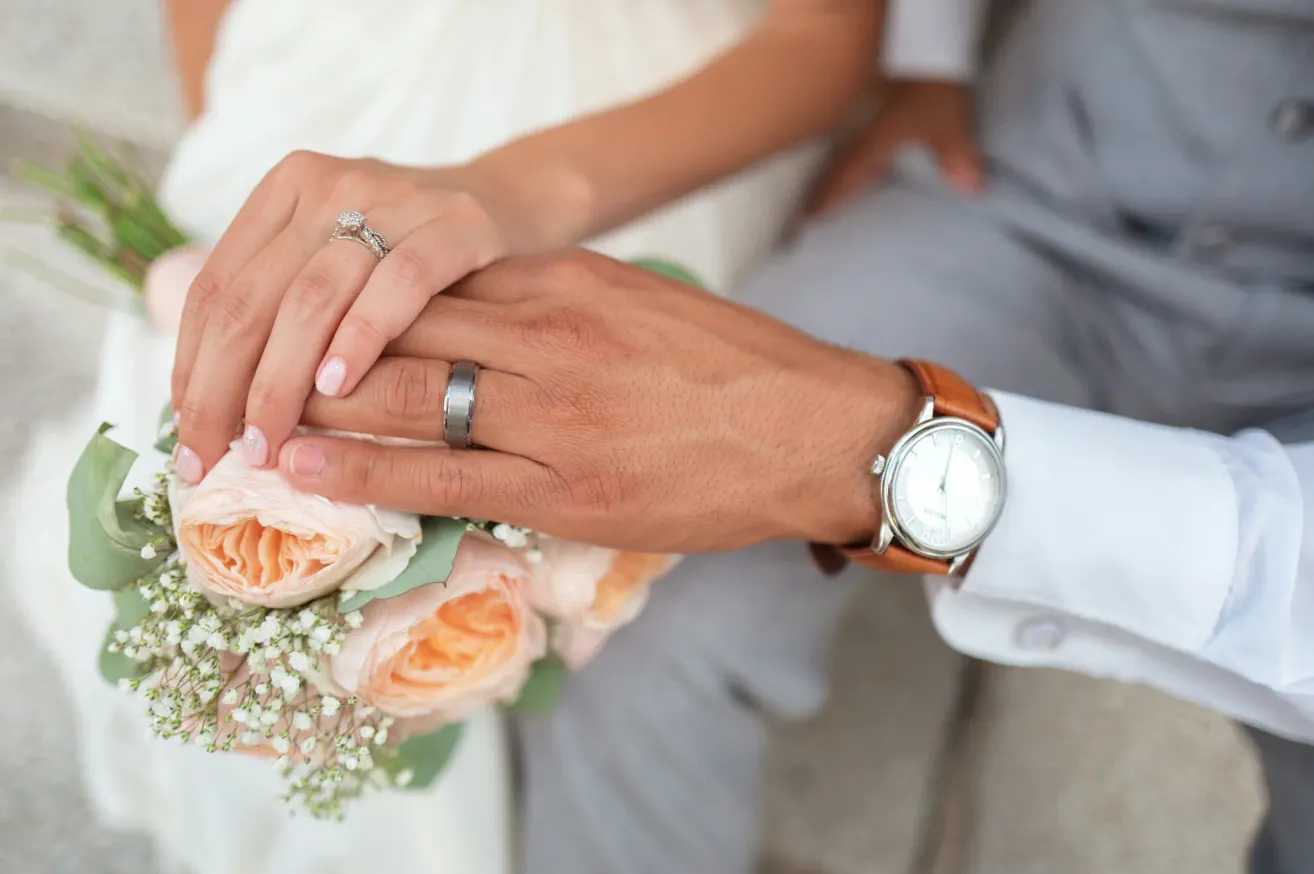 Eine Braut hält die Hand ihres Bräutigams mit einem Ring am Finger und einem Rosenstrauß. Er trägt einen Ehering und eine Uhr.