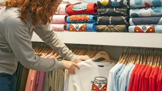 Eine Frau betrachtet ein weißes Shirt mit VW-Bus-Grafik in einem Bekleidungsgeschäft. Verschiedene bunte Shirts hängen hinter ihr an Kleiderbügeln.