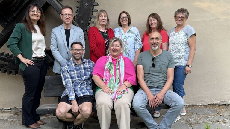 Eine Gruppe von Menschen, darunter Männer und Frauen, lächelt und posiert für ein Foto vor einer Kulisse.