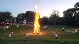 Ein Lagerfeuer brennt hell in einem Grasfeld, umgeben von Bäumen. Kinder stehen um es herum und halten brennende Fackeln, einige sind weiter weg. Der Himmel ist klar und es gibt Gebäude im Hintergrund.