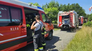 Ein Mann und ein Junge in Feuerwehruniformen stehen vor einem roten Feuerwehrwagen. Der Wagen ist am Straßenrand geparkt. Im Hintergrund sind Bäume und Büsche.