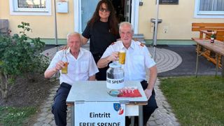 Drei Menschen sitzen an einem Tisch, lächeln und posieren für ein Foto. Ein Mann hält ein Glas Bier, der andere eine Flasche. Eine Frau steht hinter ihnen, und ein Glas mit Geld steht auf dem Tisch.