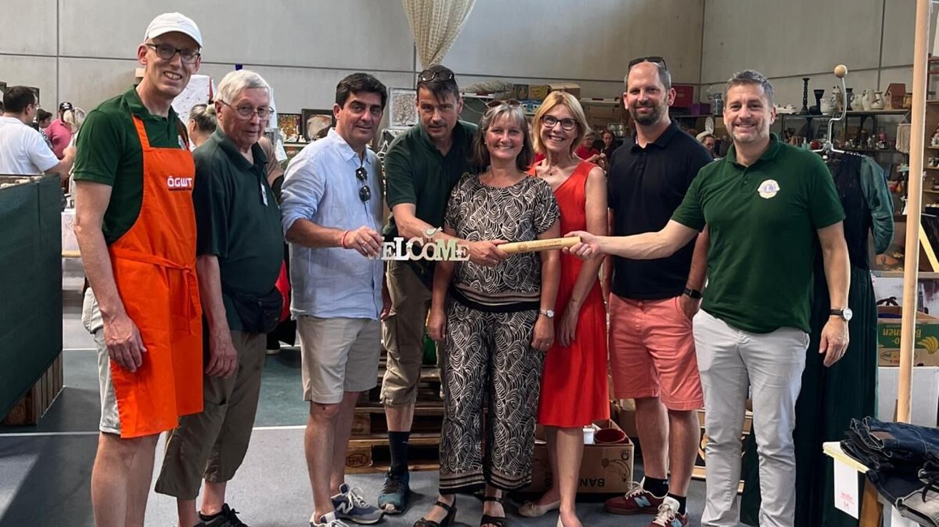 Eine Gruppe von Erwachsenen, die lächeln, stehen um ein Schild herum, das 'WELCOME' steht. Sie halten einen Holzstab, wahrscheinlich für ein Foto. Sie tragen lässige Kleidung und stehen auf einem Betonboden.