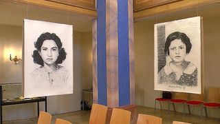 Zwei Porträts hängen von der Decke in einem Raum mit Holzstühlen, einem Schreibtisch und einer blauen Säule. Jedes Porträt zeigt eine Frau, eine mit einem ernsten Gesichtsausdruck und die andere mit einem nachdenklichen Blick.