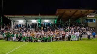 Eine große Menschenmenge in grünen und weißen Trikots steht nachts auf einem Feld, posiert für ein Gruppenfoto. Einige halten Pokale. Das Feld ist gut beleuchtet.