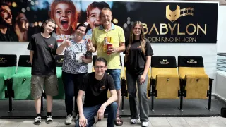 Fünf Personen posieren für ein Foto in einem Kino mit gelben Stühlen und einem großen Plakat hinter ihnen. Sie alle halten Getränke in den Händen.