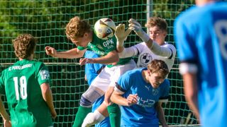 Fußballspieler kollidieren während eines Spiels. Ein Spieler in Grün versucht, den Ball zu köpfen, während ein anderer in Weiß versucht, ihn zu blockieren. Ein dritter Spieler in Blau schaut zu.