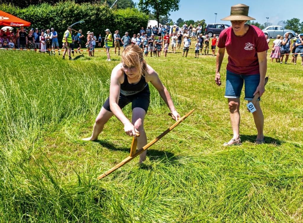 Eine Frau nimmt an einer traditionellen Aktivität teil, wobei sie ein hölzerne Werkzeug benutzt, mit Zuschauern hinter einer Absperrung auf einem Rasen.