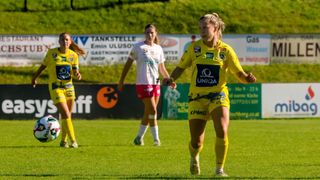 Drei weibliche Fußballspielerinnen auf einem Feld, zwei in Gelb und eine in Weiß. Sie sind in Aktion, mit grünem Rasen im Hintergrund. Banner mit verschiedenen Logos und Texten sind im Hintergrund.