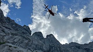 Ein Rettungshubschrauber mit einem Kreuz am Heck fliegt über felsige Berge. Er zieht jemanden in einem Gurt mit einem Seil.
