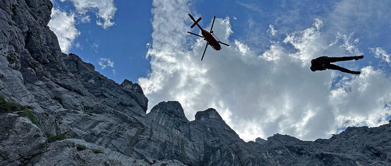 Ein Rettungshubschrauber mit einem Kreuz am Heck fliegt über felsige Berge. Er zieht jemanden in einem Gurt mit einem Seil.