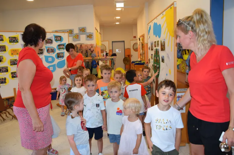 Eine Gruppe junger Kinder in einem Flur, mit einer Frau, die vor ihnen steht, lächelt und die Kinder ansieht.