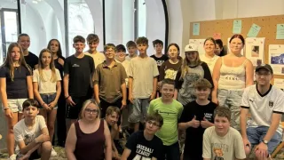 Eine Gruppe von Schülern und Lehrern posiert für ein Foto in einem Raum mit Rundbogenfenstern und einer Pinnwand. Einige Schüler tragen Brillen und Turnschuhe.