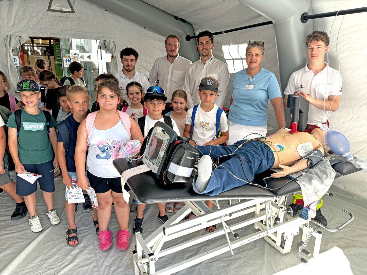 Eine Gruppe von Kindern und Erwachsenen in einem Zelt versammelt sich um einen medizinischen Mannequin auf einer Trage, einige tragen Hüte und Brillen. Der Mannequin hat Drähte und einen Monitor angeschlossen.