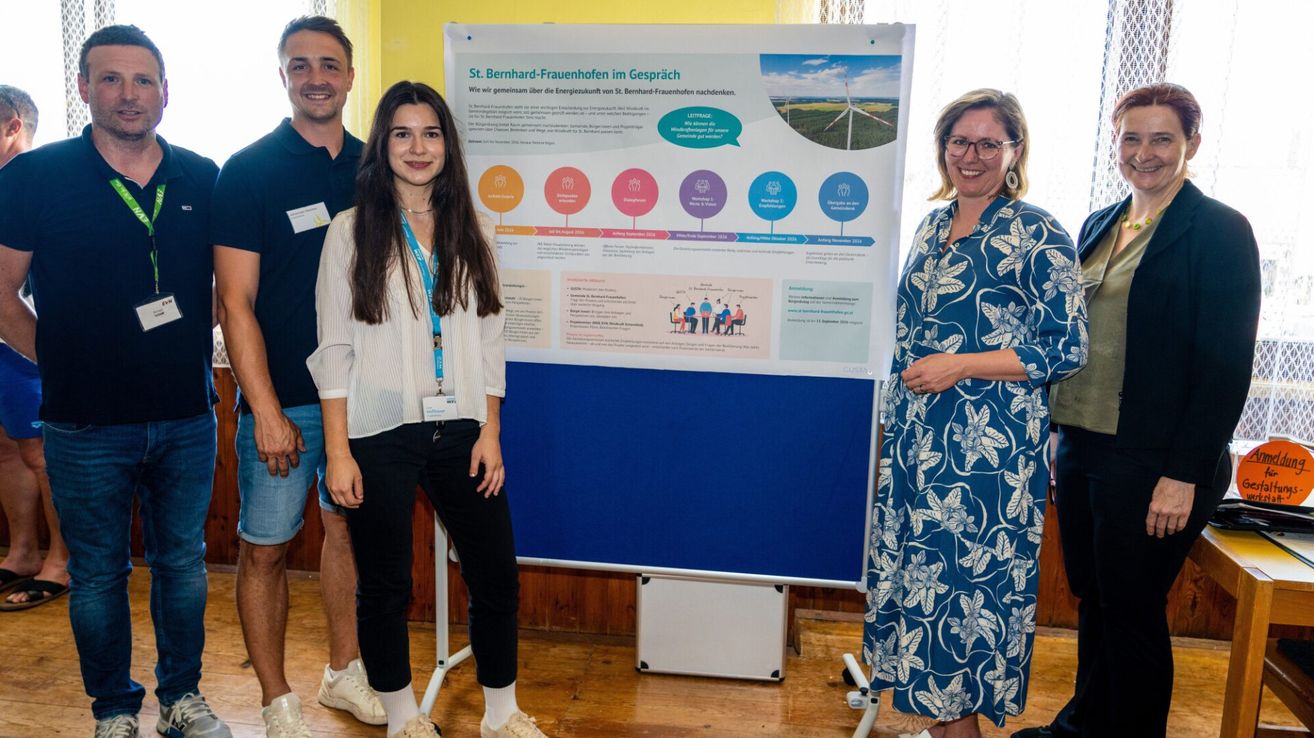 Drei Personen stehen vor einem Poster mit dem Titel 'St. Bernhard-Frauenhofen im Gespräch', wobei eine Frau in der Mitte mit einer blauen Ausweiskarte steht.