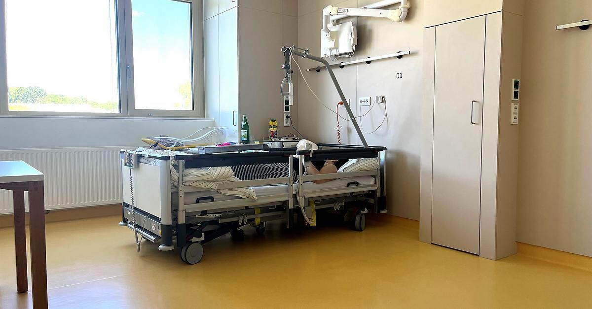 Krankenzimmer mit einem Patienten, der auf einem Bett liegt. Ausrüstung und medizinische Geräte sind am Bett befestigt. Der Raum hat ein Fenster und einen gelben Boden.