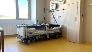 Krankenzimmer mit einem Patienten, der auf einem Bett liegt. Ausrüstung und medizinische Geräte sind am Bett befestigt. Der Raum hat ein Fenster und einen gelben Boden.