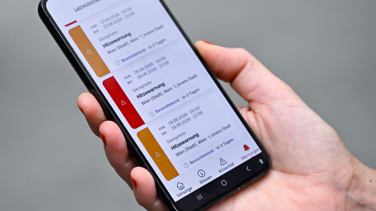 Eine Hand hält ein Smartphone mit Wetterwarnungen. Die Warnungen umfassen Hitze- und Starkregenwarnungen in Wien.