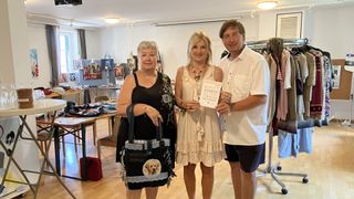 Drei Personen stehen in einem Raum mit weißer Wand und Holzboden. Eine Frau hält eine Tasche mit einem Hundebild, und ein Mann hält ein Papier. Sie stehen neben einer Frau, die eine Halskette trägt.