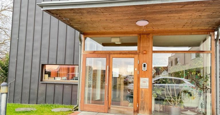 Außenansicht eines modernen Gebäudeeingangs mit Glastüren, einem Holzrahmen und einer Leuchte darüber. Ein Auto ist draußen geparkt und reflektiert sich im Glas.