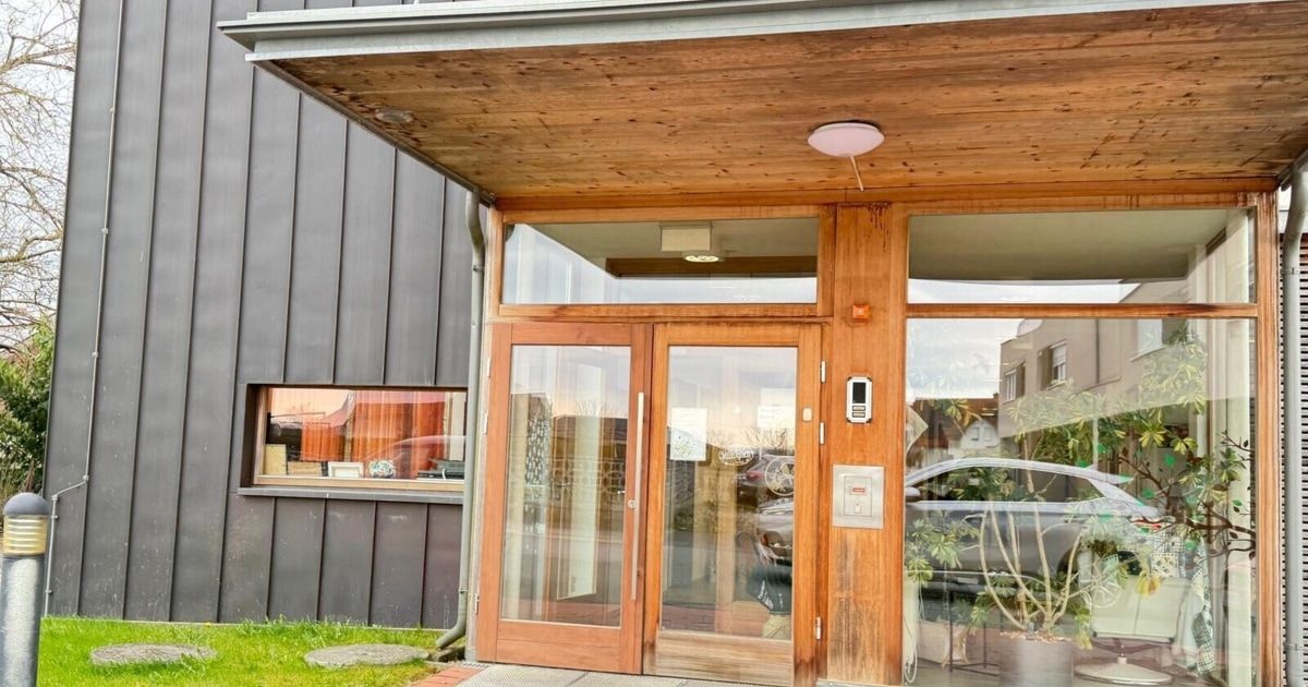 Außenansicht eines modernen Gebäudeeingangs mit Glastüren, einem Holzrahmen und einer Leuchte darüber. Ein Auto ist draußen geparkt und reflektiert sich im Glas.
