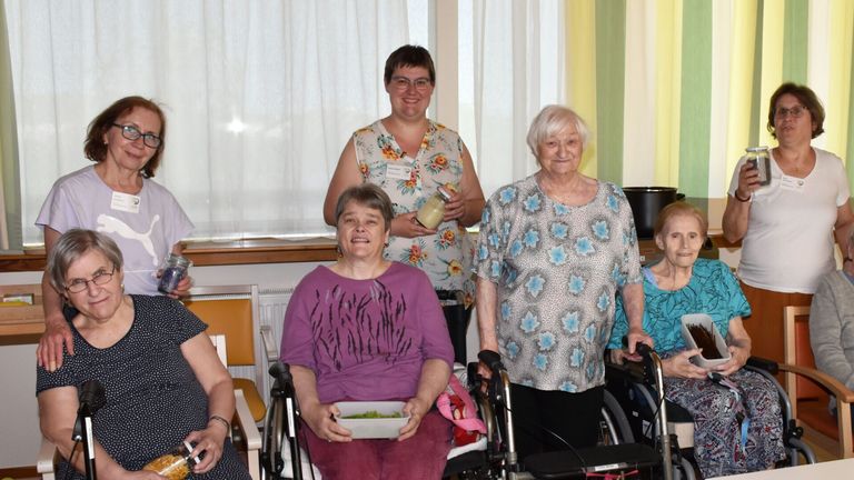 Mehrere Frauen in Rollstühlen und Stühlen posieren für ein Foto. Eine Frau in der Mitte hält eine Schüssel mit Salat. Die Frau auf der rechten Seite hält eine Flasche. Eine Frau mit Brille steht hinter ihnen. Ein Fenster mit Vorhängen ist im Hintergrund.