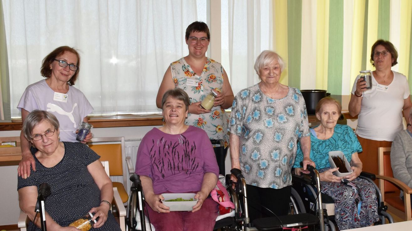 Mehrere Frauen in Rollstühlen und Stühlen posieren für ein Foto. Eine Frau in der Mitte hält eine Schüssel mit Salat. Die Frau auf der rechten Seite hält eine Flasche. Eine Frau mit Brille steht hinter ihnen. Ein Fenster mit Vorhängen ist im Hintergrund.