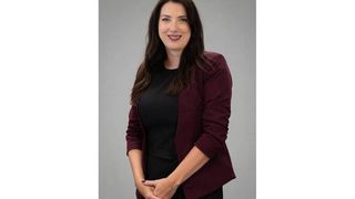 Eine Frau mit langen braunen Haaren und einem schwarzen Shirt posiert für ein Foto in einem weinroten Blazer. Sie steht vor einem grauen Hintergrund und lächelt.
