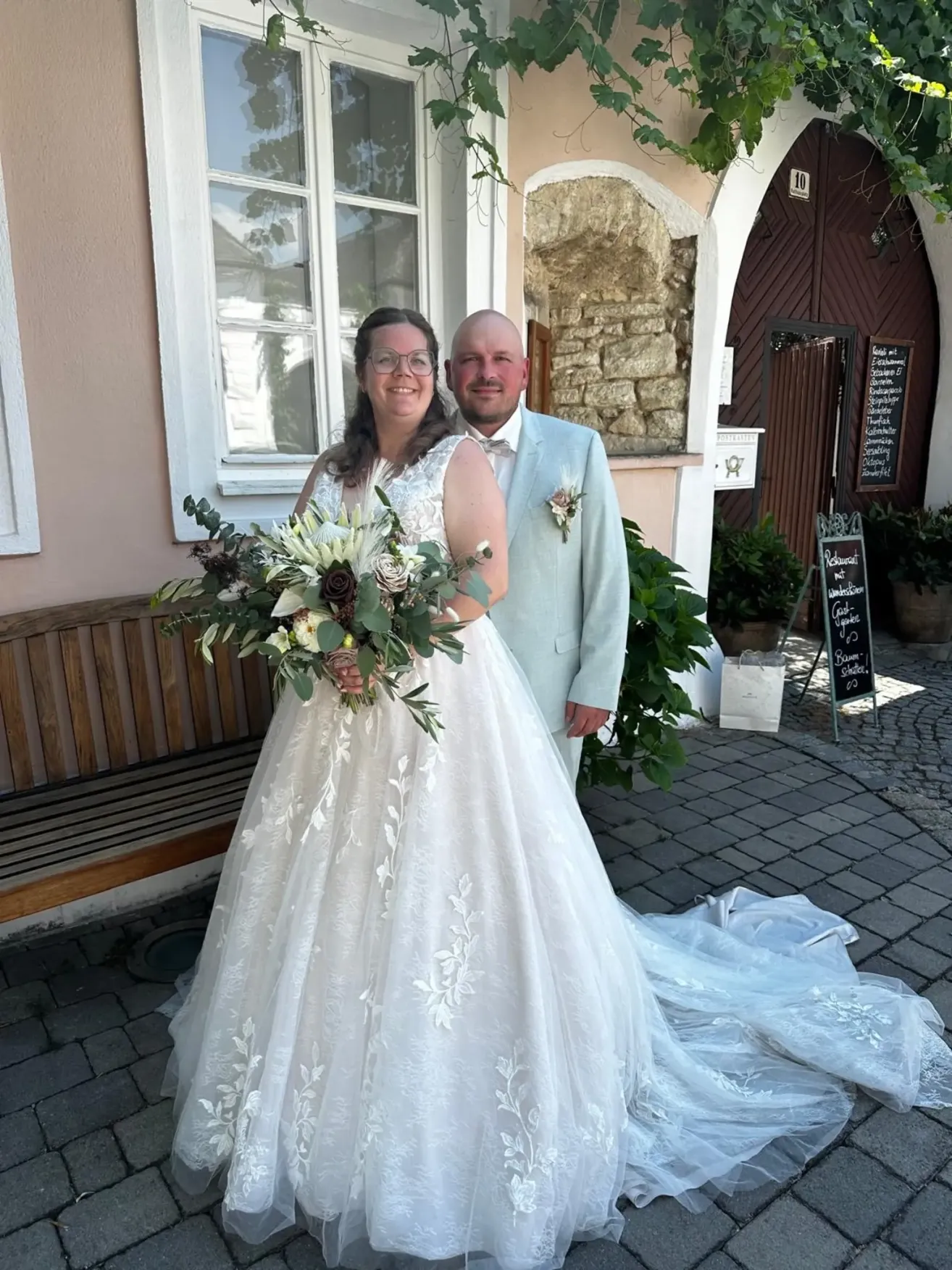 Eine lächelnde Braut in einem weißen Kleid und ein Bräutigam in einem hellen Anzug stehen vor einem Gebäude mit einer Holztür.