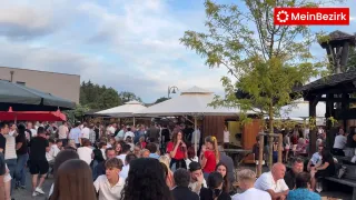 Eine große Menschenmenge versammelt sich im Freien, einige unter Schirmen, bei einer geschäftigen Outdoor-Veranstaltung. Bäume und Schirme bieten Schatten. Eine Frau in einem roten Shirt trinkt aus einer Flasche.
