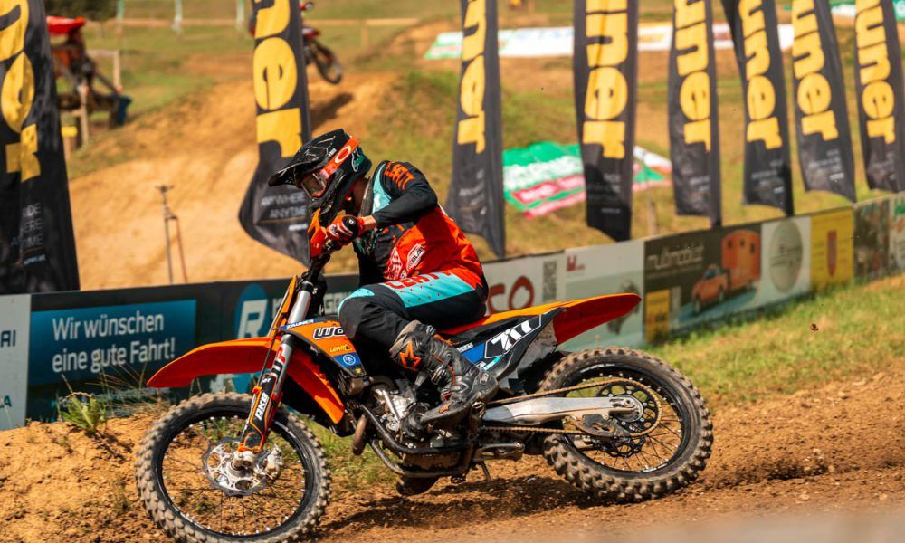 Ein Motocross-Fahrer mit Helm, Handschuhen und Rennanzug fährt ein orangefarbenes Motorrad auf einer Erdstrecke.