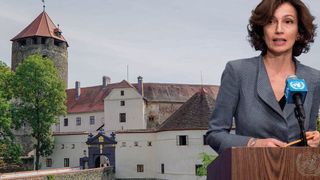 Eine Frau spricht an einem Podium vor einer Burg mit rotem Dach und vielen Fenstern.