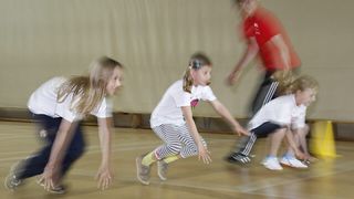 Drei Kinder spielen auf einem Holzboden, zwei in weißen Shirts, eines mit blauen Haarklemmen, das andere gebeugt. Ein drittes Kind in Rot ist verschwommen.
